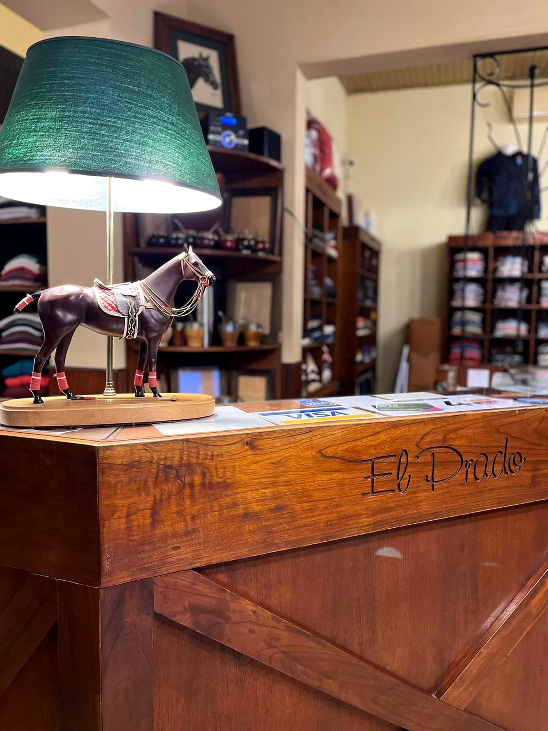 Lámpara de mesa con pantalla verde y un caballo criollo de adorno, sobre el mostrador tallado con el nombre El Prado.